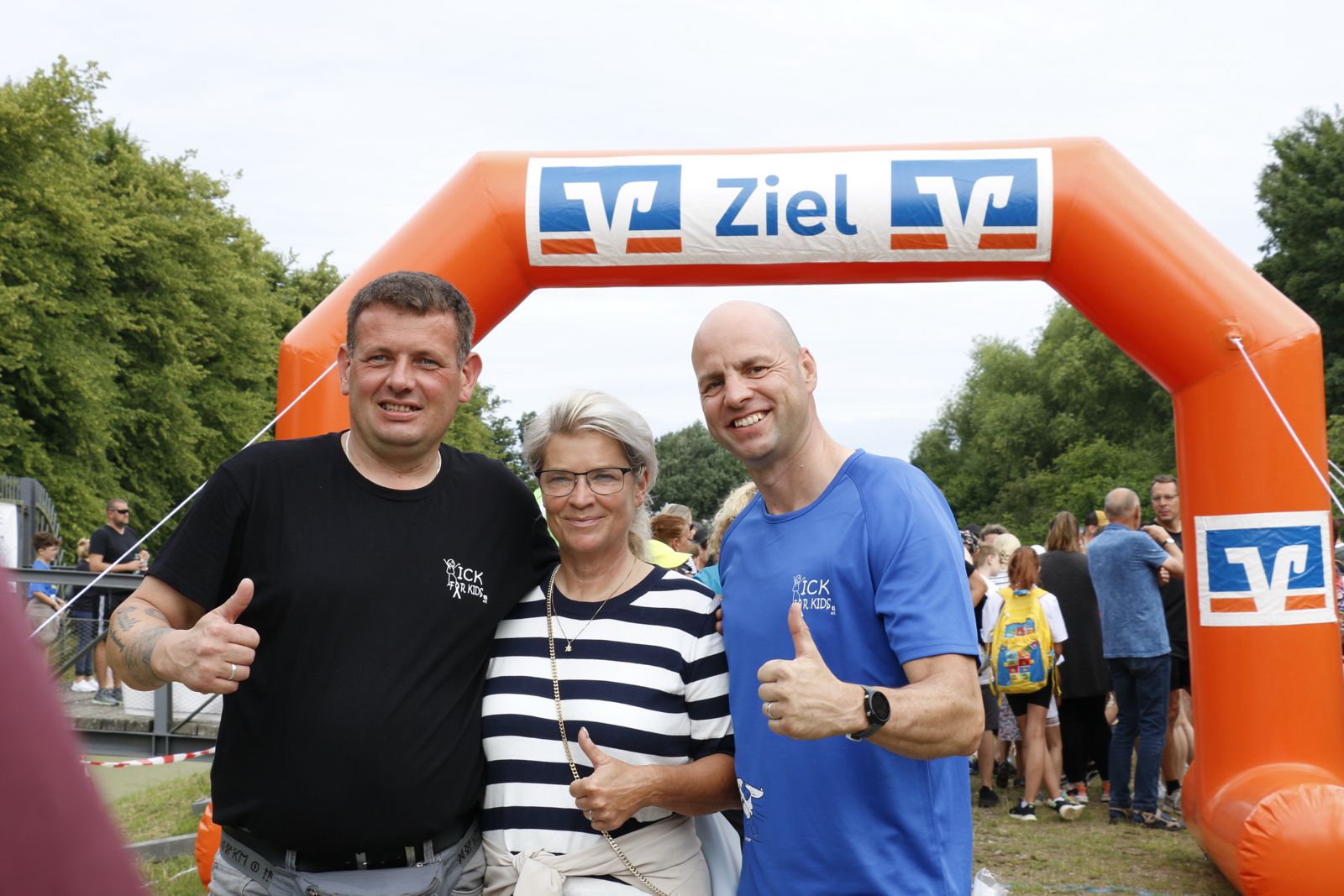 Drei Organisatoren des Kick for Kids e.V. posieren lächelnd vor dem orangefarbenen Zielbogen der VR Bank